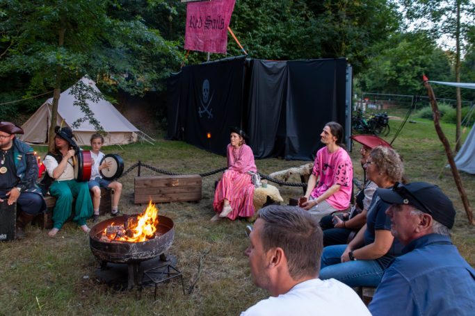 Lagerfeuer mit Leuten in Gesellschaft, umgeben von Zelten und Bäumen.