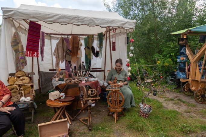 Marktstand mit Handwerk, Spinnrad und Ständen für handgemachte Waren. Menschen sind aktiv.