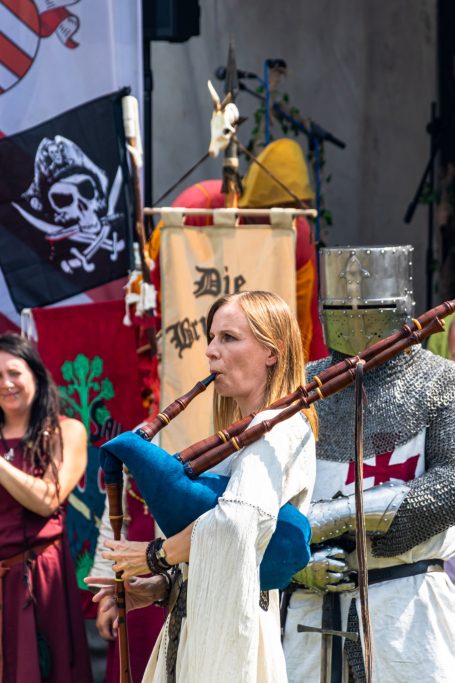 Frau in historischer Kleidung spielt Dudelsack auf einem Fest, im Hintergrund Flaggen.
