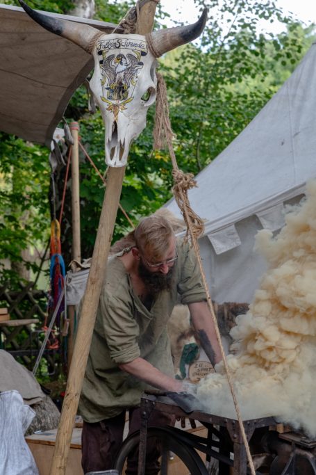 Handwerker bearbeitet Holz unter einem Zeltdach, im Hintergrund ein Tierkopf-Skelett.