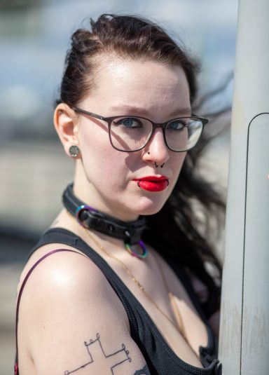 Frau mit Brille, langen Haaren, Tattoos und einem Halsband, schaut nachdenklich.