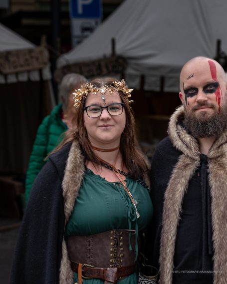 Frau in grüner Kleidung mit Kranz, Mann mit schwarzem Kriegsanstrich und Fellumhang.