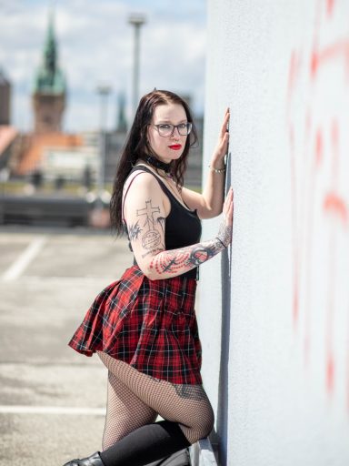Frau mit Brille und Tattoos in einem kurzen karierten Rock, posiert an einer Wand.