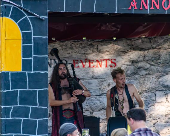 Zwei Musiker in Mittelalterkleidung spielen auf einer Bühne vor einer steinernen Wand.