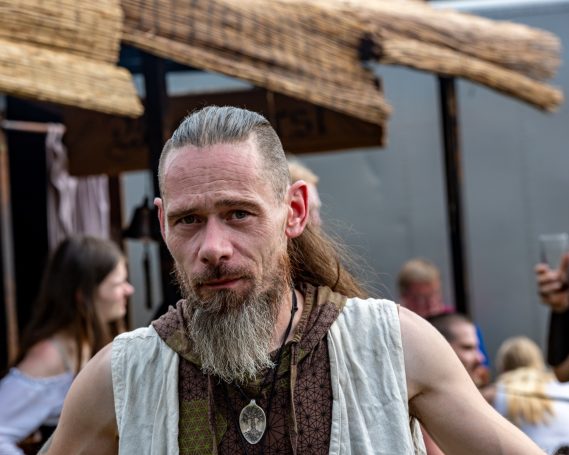 Ein Mann mit Bart und langem Haar, trägt eine traditionelle Kleidung und schaut direkt.