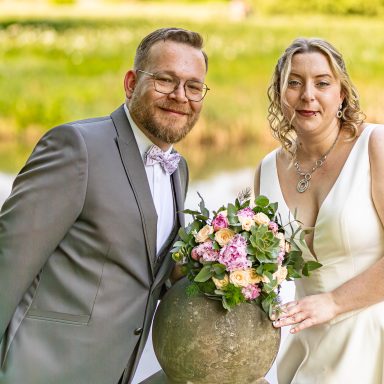 Hochzeitspaar steht am Teich mit einem Blumenstrauß in einer stilvollen Vase.