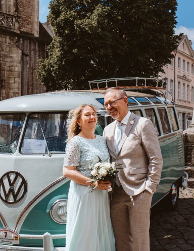 Paar in festlicher Kleidung steht vor einem retro Volkswagen-Bus, umgeben von Bäumen.