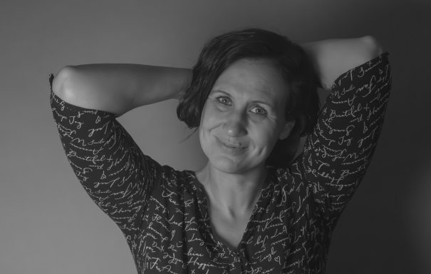 Portrait einer Frau mit lockigem Haar, die lächelt und die Arme hinter dem Kopf verschränkt.