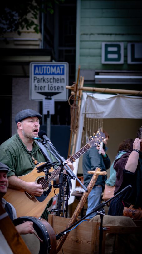 Musiker mit Gitarre singt auf einer Straße, im Hintergrund Stände und Publikum.