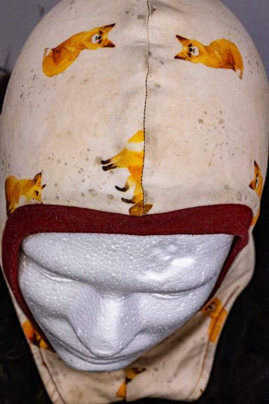 Beige Mütze mit orangefarbenen Fuchs-Motiven und rotem Rand.