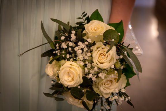 Hochzeitsstrauß mit weißen Rosen und grünen Blättern, gehalten von einer Person.