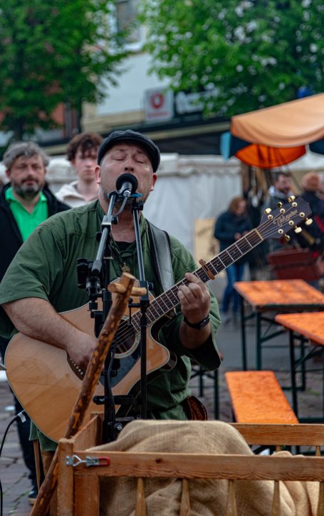 Mann mit Gitarre singt auf einem Markt, Publikum im Hintergrund.