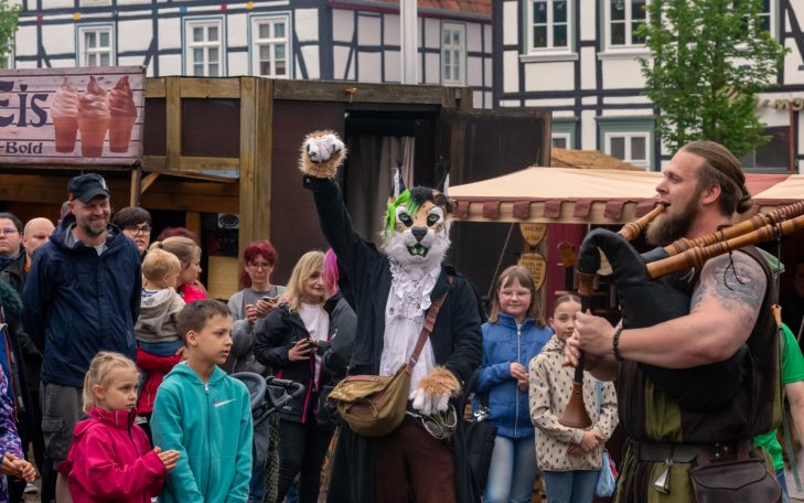 Person in Tiermaske mit einer Gruppe von Menschen, die einem Straßenkünstler zuschauen.